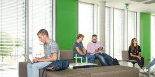 Studierende in einem Gebäude der Uni Sechs Studierende sitzen in einem Aufenthaltsbereich in einem Uni-Gebäude. Sie lesen Bücher, arbeiten am Laptop oder unterhalten sich.
