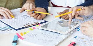 Diagramme_03 Auf einem Tisch liegen Blöcke und Blätter, ein Tablett, welches von einer Hand benutzt wird.